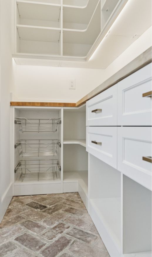 A modern, empty pantry with white shelves, wire pull-out baskets, drawers with gold handles, a wooden countertop, and a herringbone-patterned tile floor—perfect inspiration from a custom closet designer.