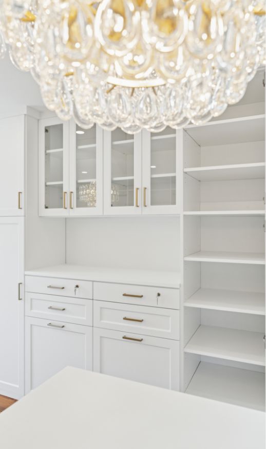 Modern white kitchen with glass-front cabinets, open shelves, and gold handles features a countertop by a Custom Closet Designer. A sparkling crystal chandelier hangs overhead, reflecting light across the bright, tidy space.