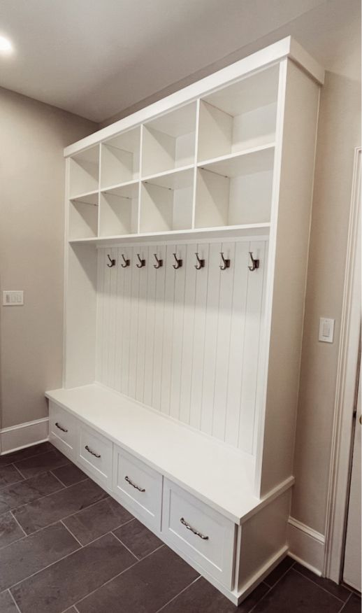 A white mudroom storage unit with open cubbies on top, a row of coat hooks in the middle, and bench seating with four pull-out drawers underneath offers custom storage solutions against a light-colored wall and tiled floor.