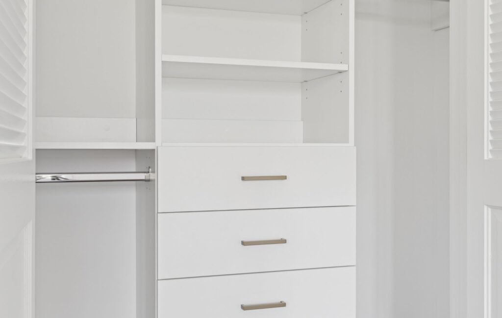 A modern white closet with three drawers, open shelves above, and a metal hanging rod on the left side, crafted by a Custom Closet Designer. The closet features clean lines and a minimalistic design for ultimate Custom Storage Solutions.