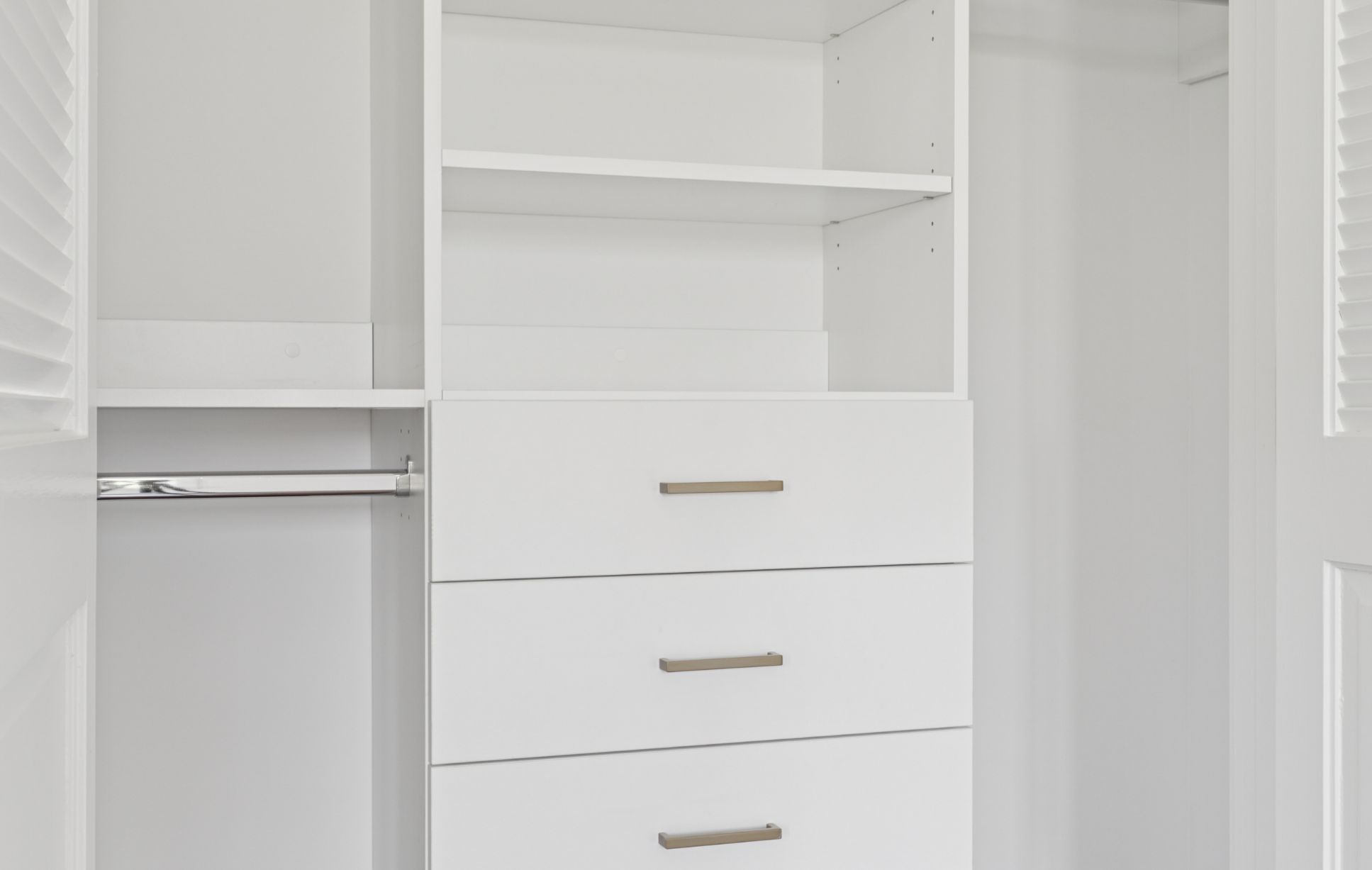 A modern white closet with three drawers, open shelves above, and a metal hanging rod on the left side, crafted by a Custom Closet Designer. The closet features clean lines and a minimalistic design for ultimate Custom Storage Solutions.