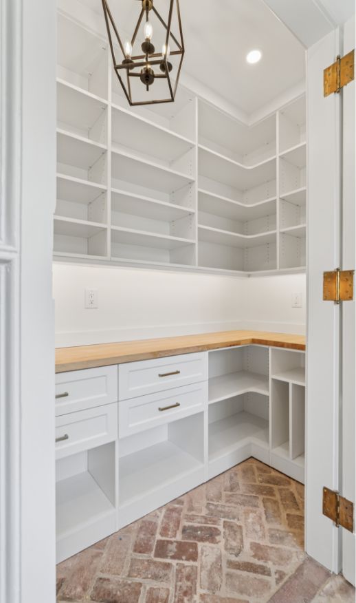 A walk-in pantry with white shelves, white drawers, wooden countertops, and a herringbone brick floor. Designed by a custom closet designer, it features a geometric pendant light and recessed lighting for added illumination.