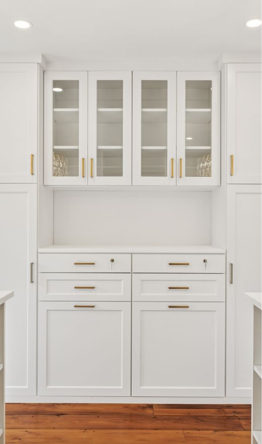 White built-in cabinets with gold handles, glass upper cabinet doors, and multiple drawers below offer elegant custom storage solutions, set against a white wall with wooden flooring. The room is well-lit with recessed ceiling lights.