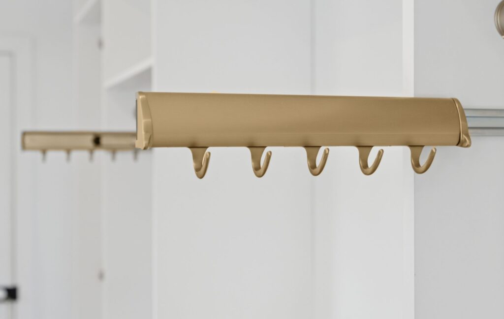 Close-up of a gold-colored metal rail with multiple hooks for hanging items, mounted inside a white closet—an elegant touch by a Custom Closet Designer. Another identical rail is visible in the background, perfect for custom storage solutions.