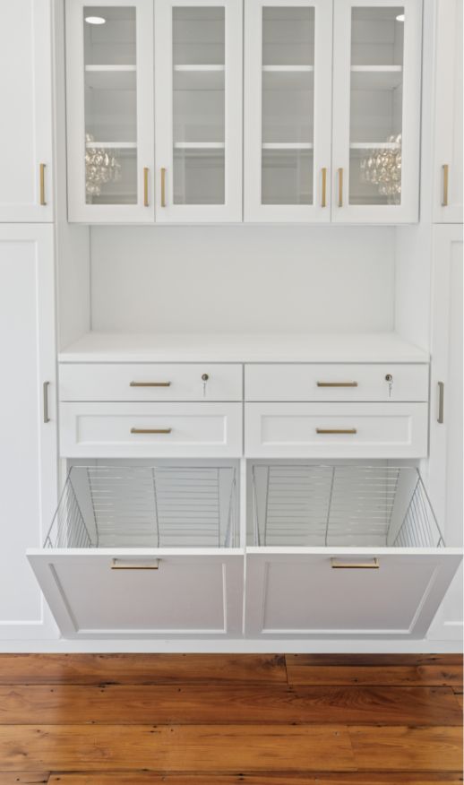 White built-in cabinetry with glass-front uppers, four drawers, and two lower compartments with pull-out wire baskets offers elegant custom storage solutions and gold handles, all beautifully set against a wooden floor.