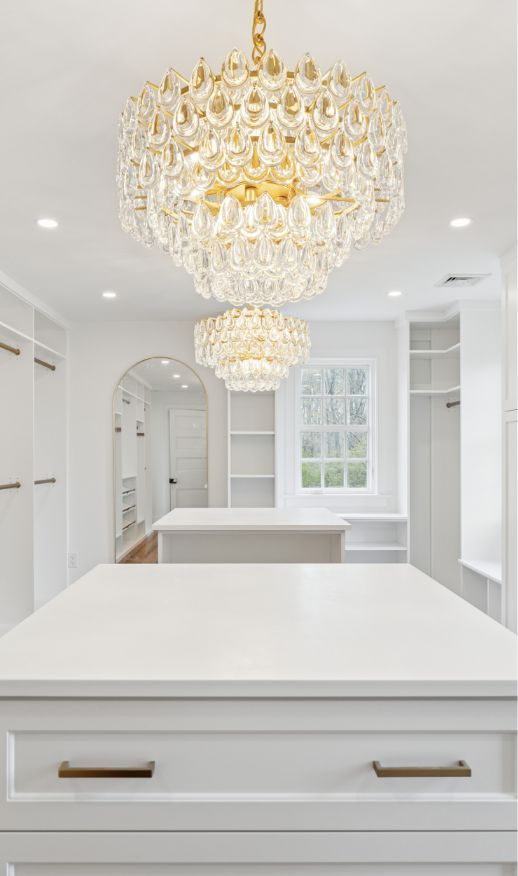 Spacious white custom walk-in closet with built-in shelves, two crystal chandeliers, a large window, an arched mirror, and a central island with gold handles—expertly crafted by a skilled custom closet designer.