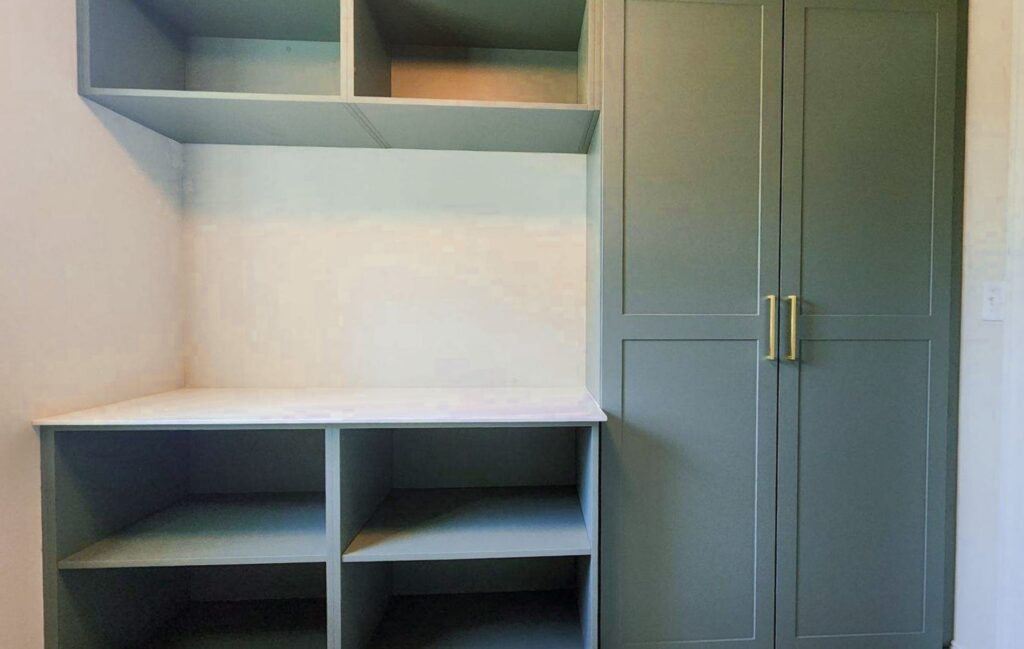 Designed by a Custom Closet Designer, these built-in shelving and cabinets feature a light grayish-green finish with open shelves on the left and two tall cabinet doors with gold handles on the right, perfect for elegant custom closets.