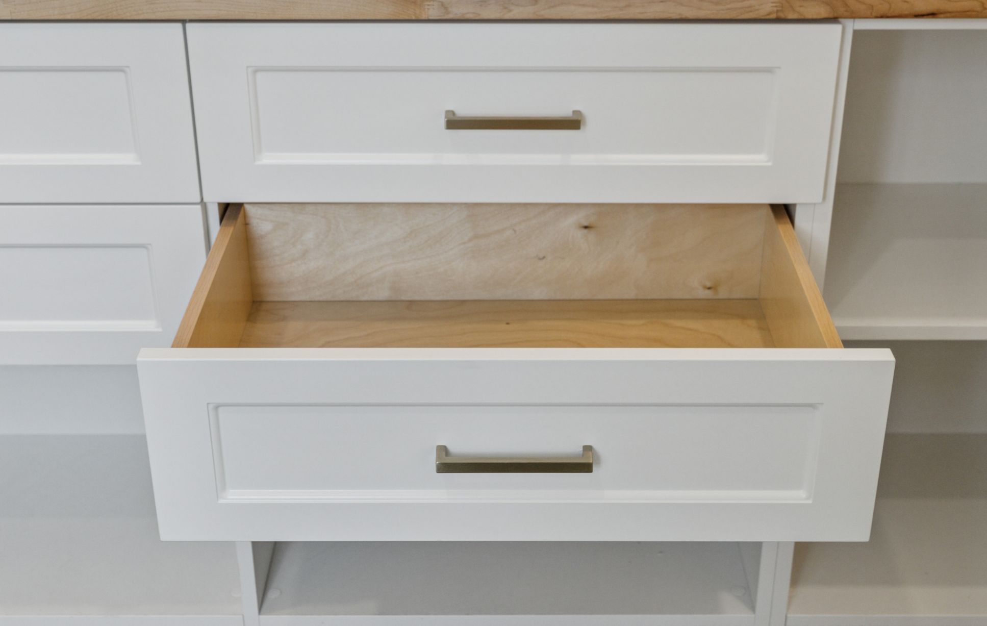 A white kitchen cabinet with an open wooden drawer, showcasing its empty interior. Featuring a metallic handle and wooden countertop, this setup exemplifies the elegance of custom storage solutions.