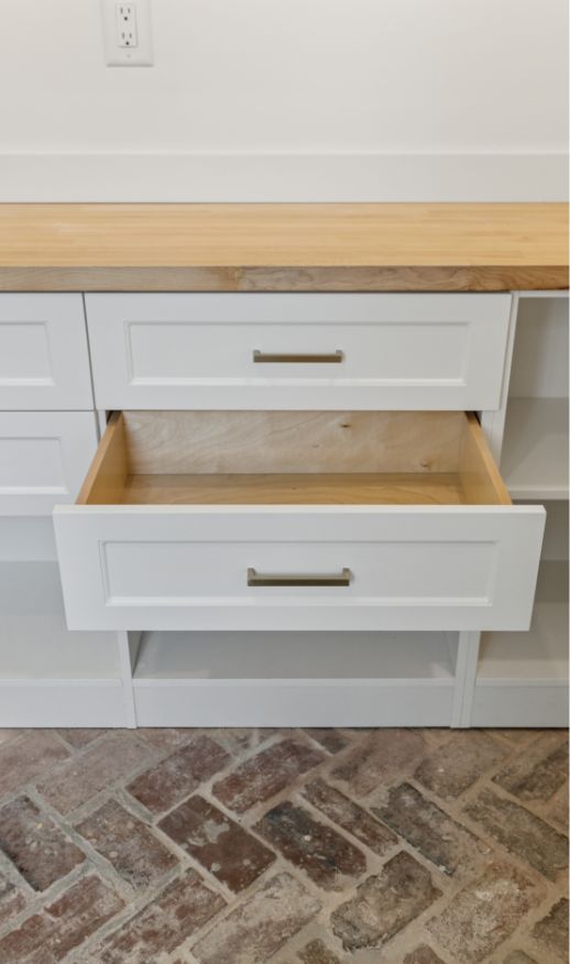 A white kitchen cabinet with a wooden countertop and brick floor showcases custom storage solutions. One large drawer is open, revealing its spacious, empty interior. The silver handles and modern design offer both style and functionality.