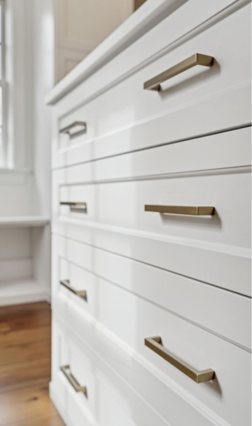 Close-up of white dresser drawers with modern, rectangular gold handles—an elegant touch from a Custom Closet Designer. The surface is clean and smooth, with a window and wooden floor visible in the background.