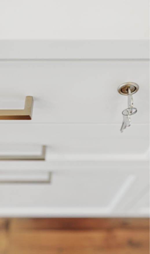 A close-up of a white cabinet drawer with a gold handle highlights elegant custom storage solutions. A key is inserted into the lock above the handle, and part of a wooden floor is visible at the bottom of the image.