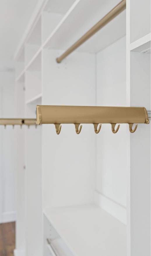 A close-up of a modern custom closet with white shelves and a gold pull-out rack featuring five hooks for hanging items. The background showcases more shelves and hanging rods—perfect for tailored custom storage solutions.