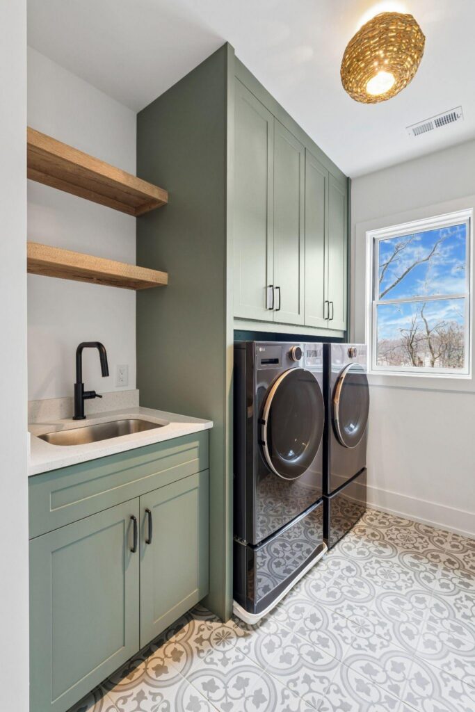Laundry room with green cabinets, black washer and dryer, patterned tile floor, a sink with black faucet, wooden shelves, and a window showing a clear sky and bare tree branches.