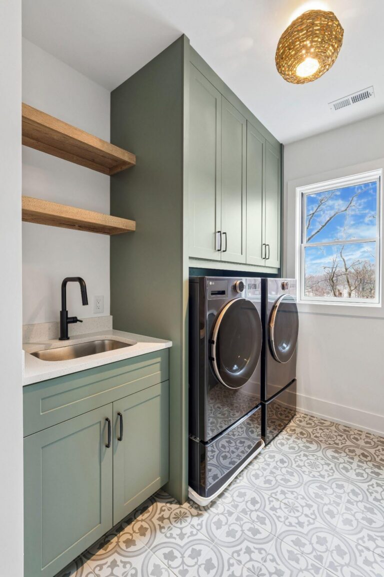Laundry room with green cabinets, black washer and dryer, patterned tile floor, a sink with black faucet, wooden shelves, and a window showing a clear sky and bare tree branches.