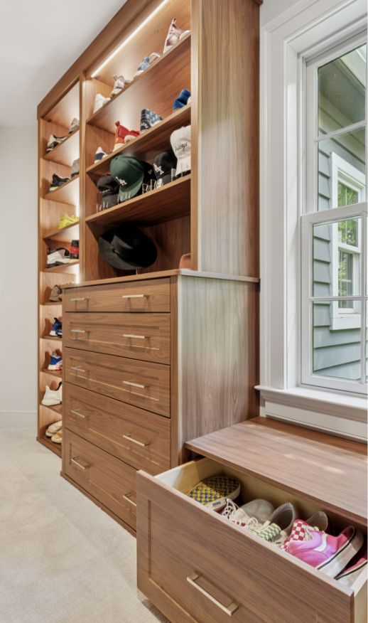 A modern wooden closet features open shelves displaying hats and shoes, a set of drawers, and a pull-out drawer beneath a window seat that holds more shoes. Natural light streams in from a nearby window.