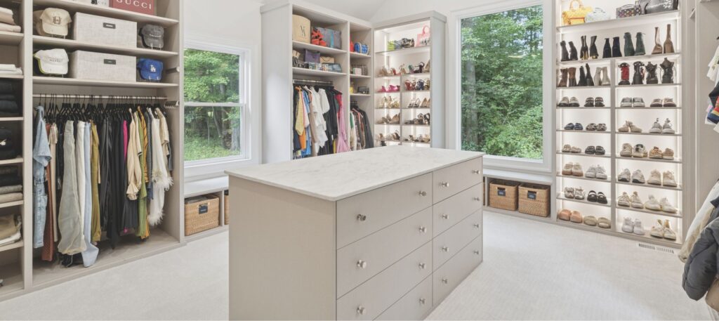 Spacious, modern walk-in closet with shelves of shoes, handbags, hats, hanging clothes, and a large island with drawers in the center. Two windows provide natural light and reveal a green, wooded view outside.
