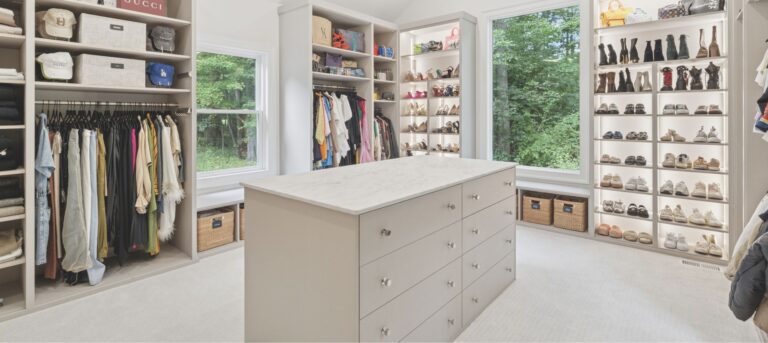 Spacious, modern walk-in closet with shelves of shoes, handbags, hats, hanging clothes, and a large island with drawers in the center. Two windows provide natural light and reveal a green, wooded view outside.