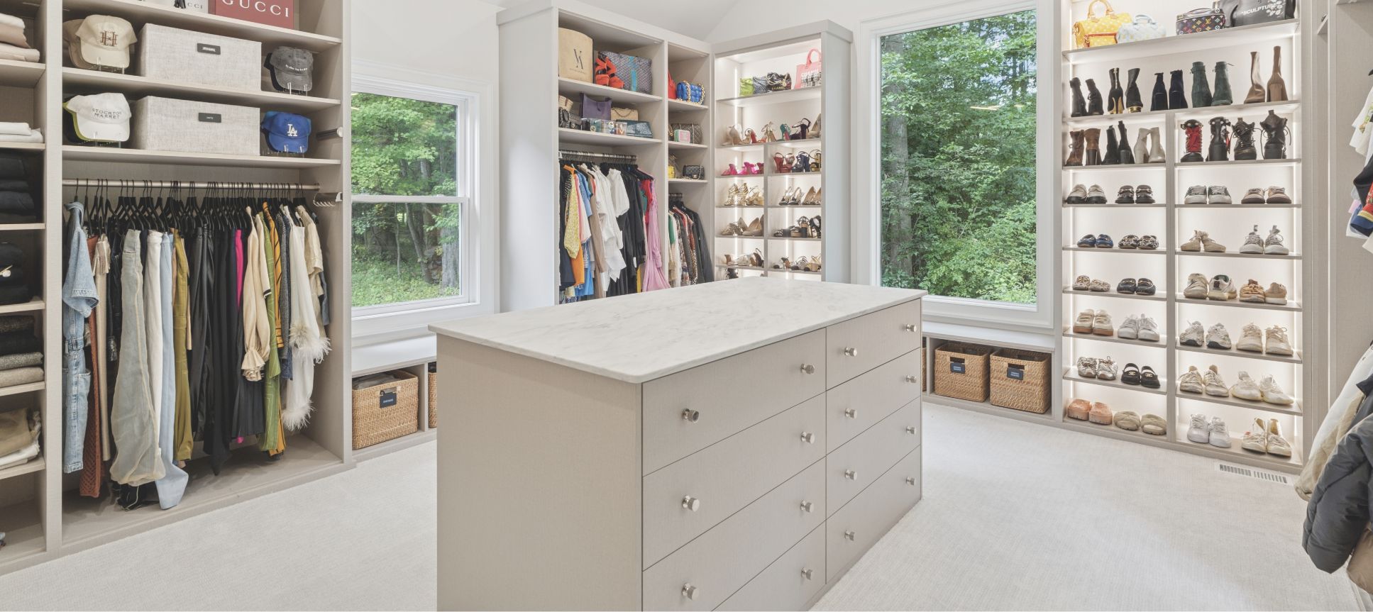 Spacious, modern walk-in closet with shelves of shoes, handbags, hats, hanging clothes, and a large island with drawers in the center. Two windows provide natural light and reveal a green, wooded view outside.
