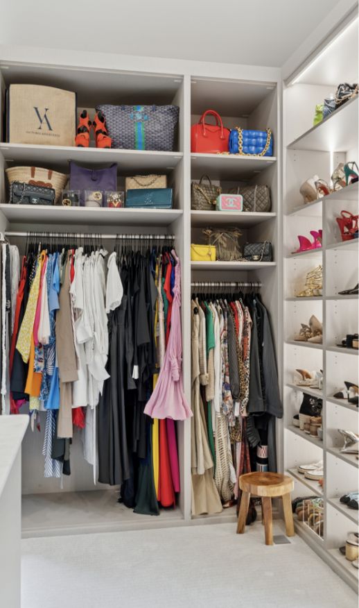 A spacious walk-in closet with neatly organized clothes hanging on racks, shelves of designer handbags and hats, cubbies filled with shoes, and a small wooden stool on the floor.