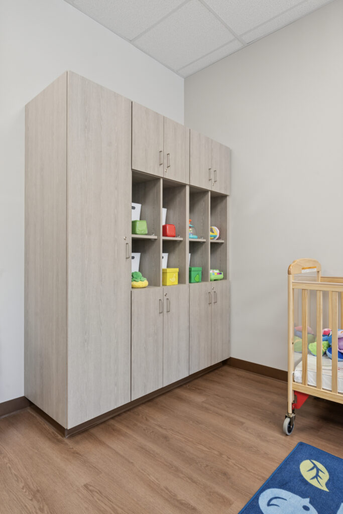 A light wood storage cabinet with open cubbies and closed doors stands against a beige wall in a room with wood flooring. Colorful toys and bins are on the shelves, and part of a crib and blue rug are visible on the right.