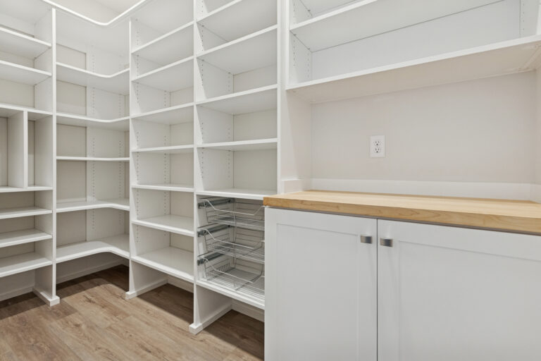 A spacious walk-in pantry with white shelves, empty wire baskets, a wooden countertop, and white cabinets. The floor is light wood, and theres an electrical outlet above the counter. Kitchen pantry storage