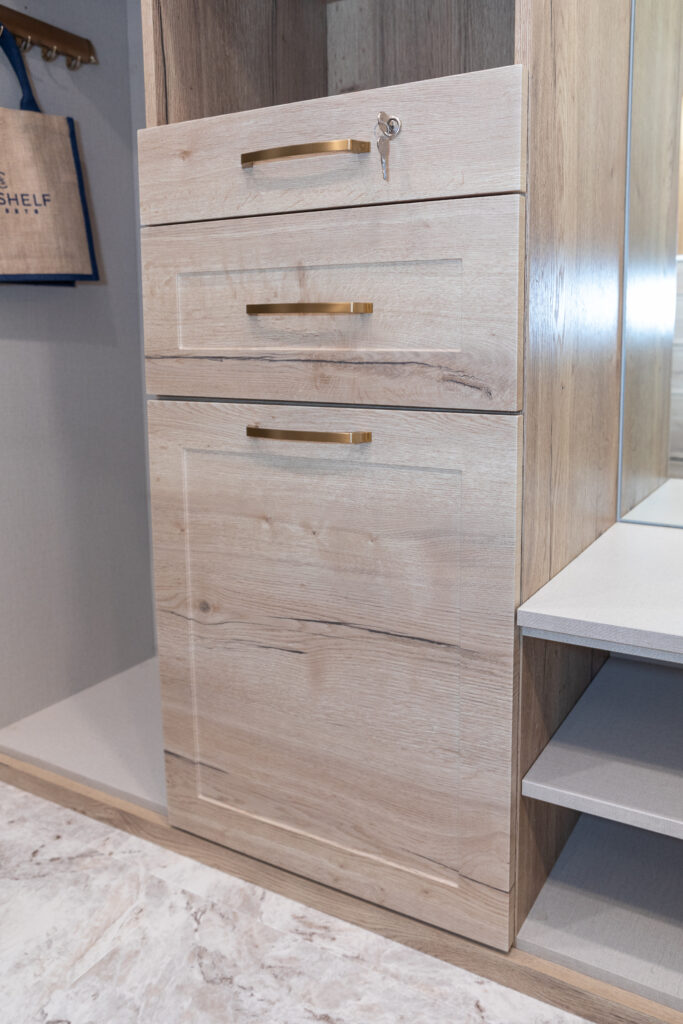 A light wood cabinet with two drawers and a compartment door, each with brass handles; the top drawer is locked with a small key. The cabinet stands on a marble-patterned floor next to a shelf and a mirror.