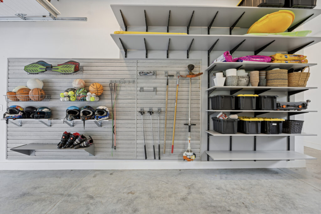 A well-organized garage wall with shelves and hooks holding sports equipment, balls, helmets, tools, baskets, and storage bins; the floor is clean and the items are neatly arranged.