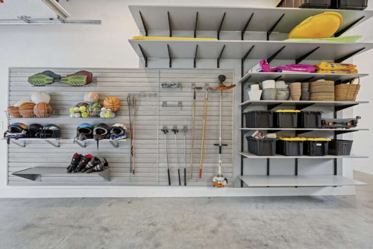A well-organized garage wall with shelves and hooks holding sports equipment, balls, helmets, tools, baskets, and storage bins; the floor is clean and the items are neatly arranged.