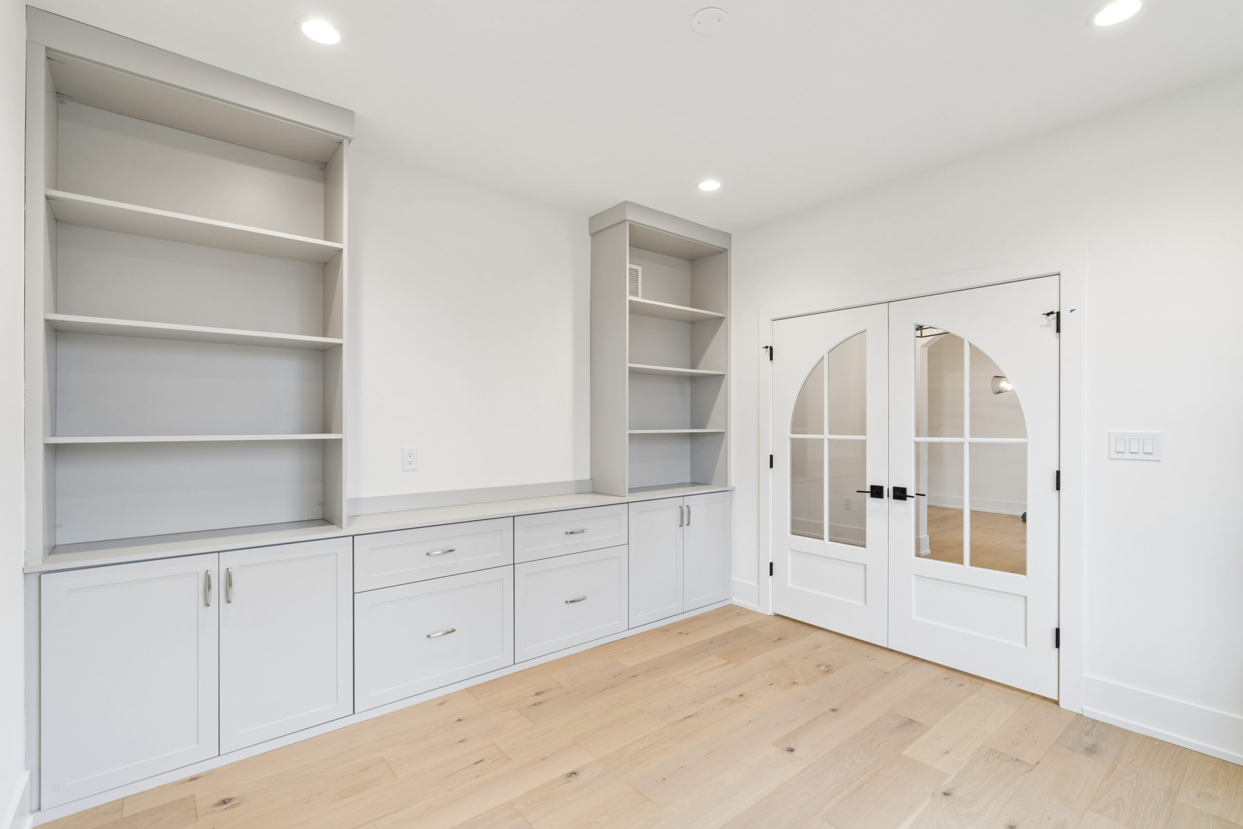 Built-in light gray cabinets with open shelves and drawers line a white wall in a bright room with light wood floors and glass double doors.