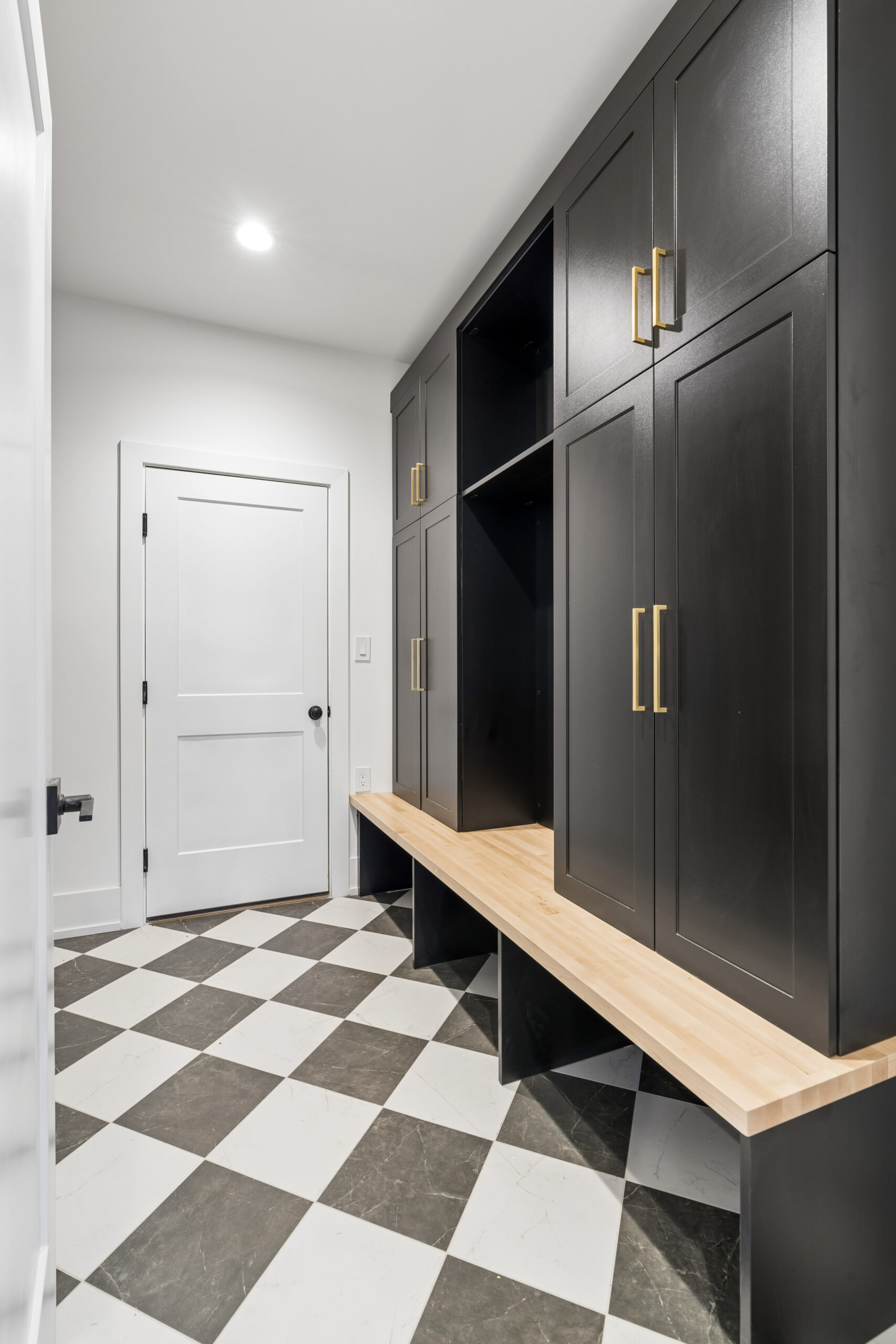 A modern mudroom with black cabinets, gold handles, a wooden bench, and black-and-white checkered tile flooring. A white door is at the end of the room.