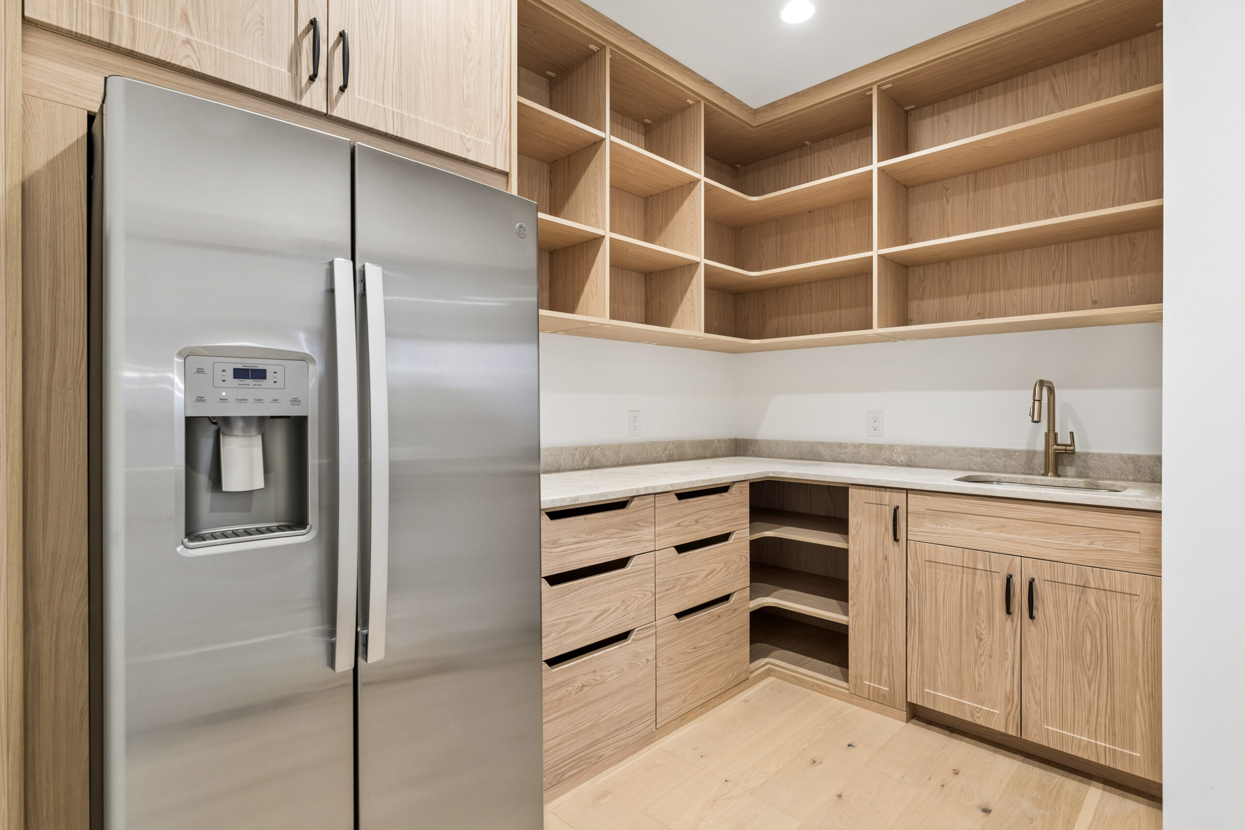 Modern kitchen pantry with light wood cabinets and open shelving, a stainless steel refrigerator with water dispenser, and a small sink with a gold faucet set in a light-colored countertop.