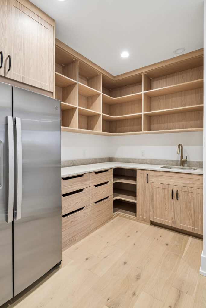 Modern kitchen pantry with light wood cabinets and shelves, stainless steel refrigerator, small sink with gold faucet, and light wood flooring. The countertops are light-colored, and there is recessed ceiling lighting.