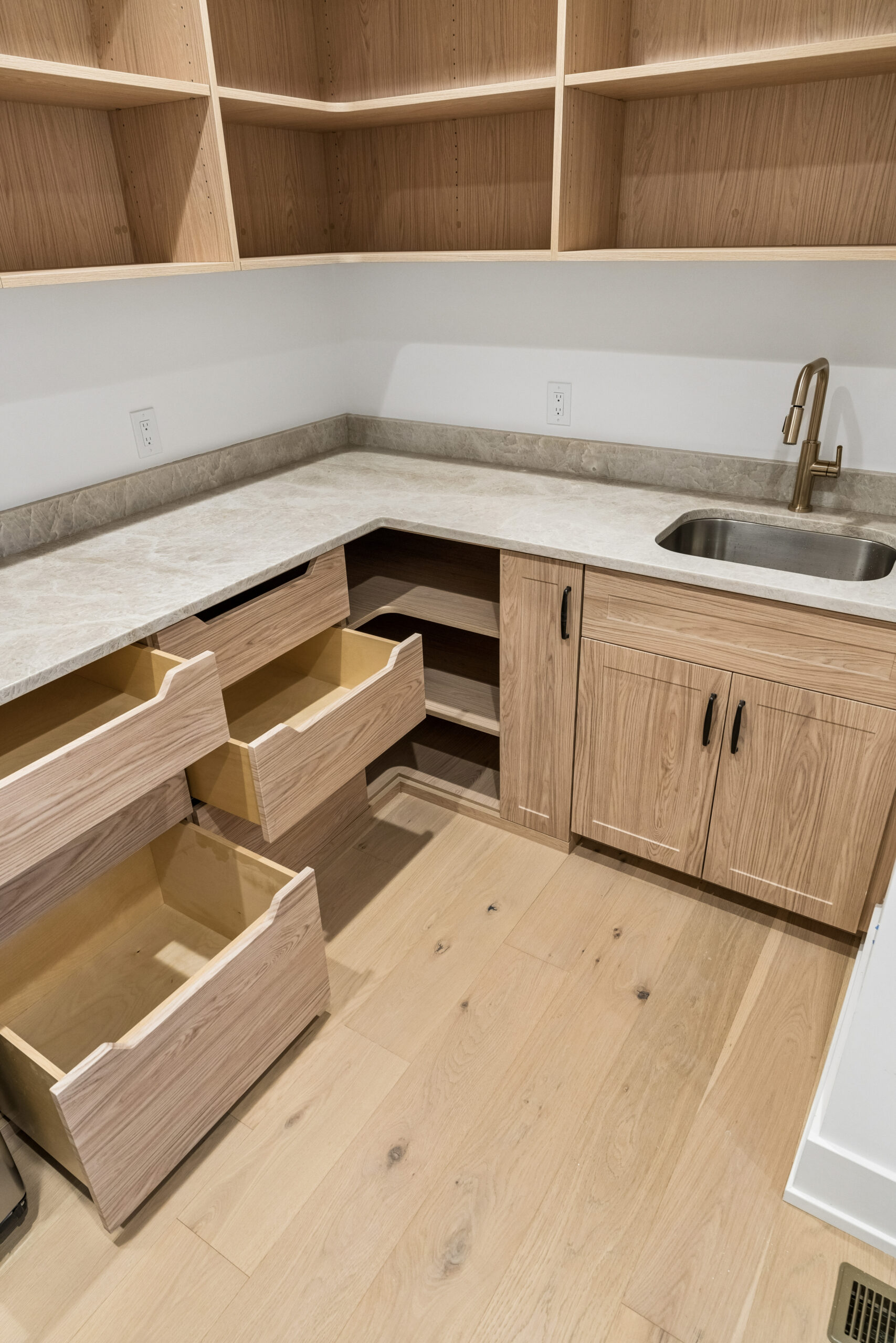 A modern kitchen area with light wood cabinets, some drawers open, open shelving above, a stone countertop, a stainless steel sink with a gold faucet, and light wood flooring.