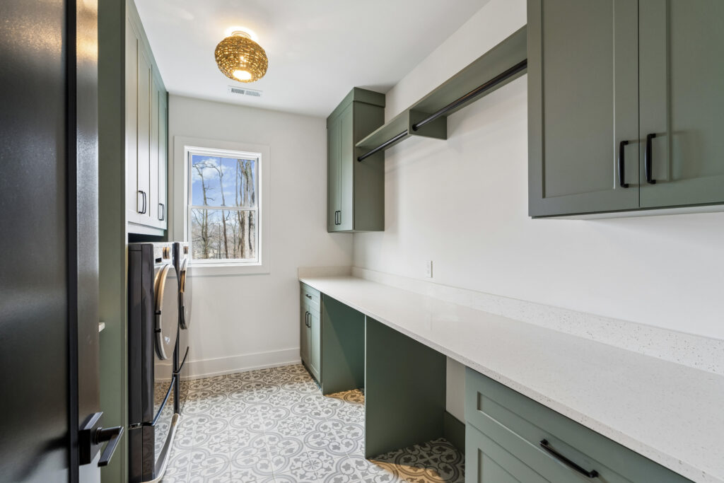 A modern laundry room with green cabinets, a long white countertop, patterned tile floor, washer and dryer by the window, and a round ceiling light fixture.