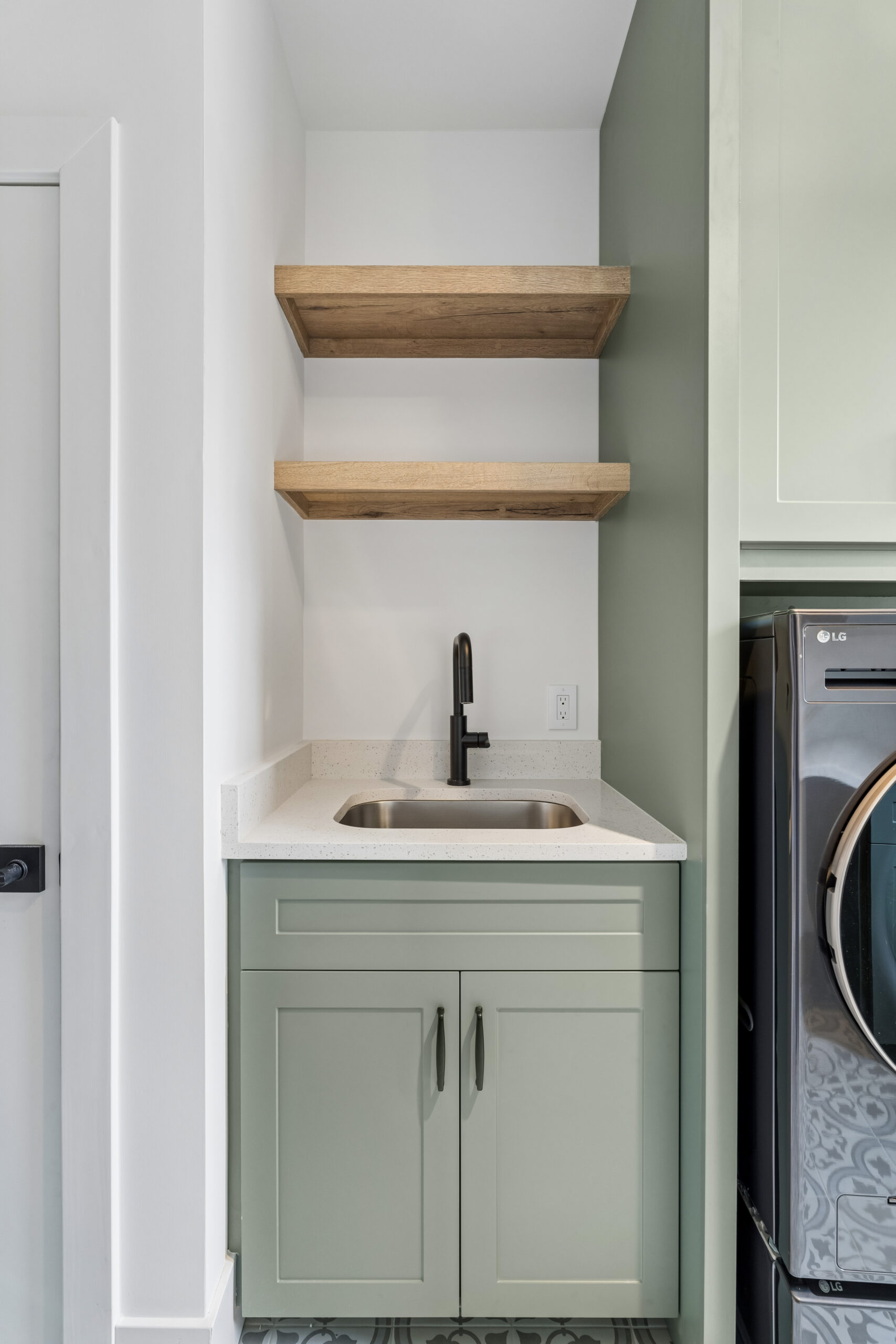 A small laundry room sink area with sage green cabinets, a white countertop, a stainless steel sink with a black faucet, two wooden floating shelves above, and part of a washing machine visible on the right.