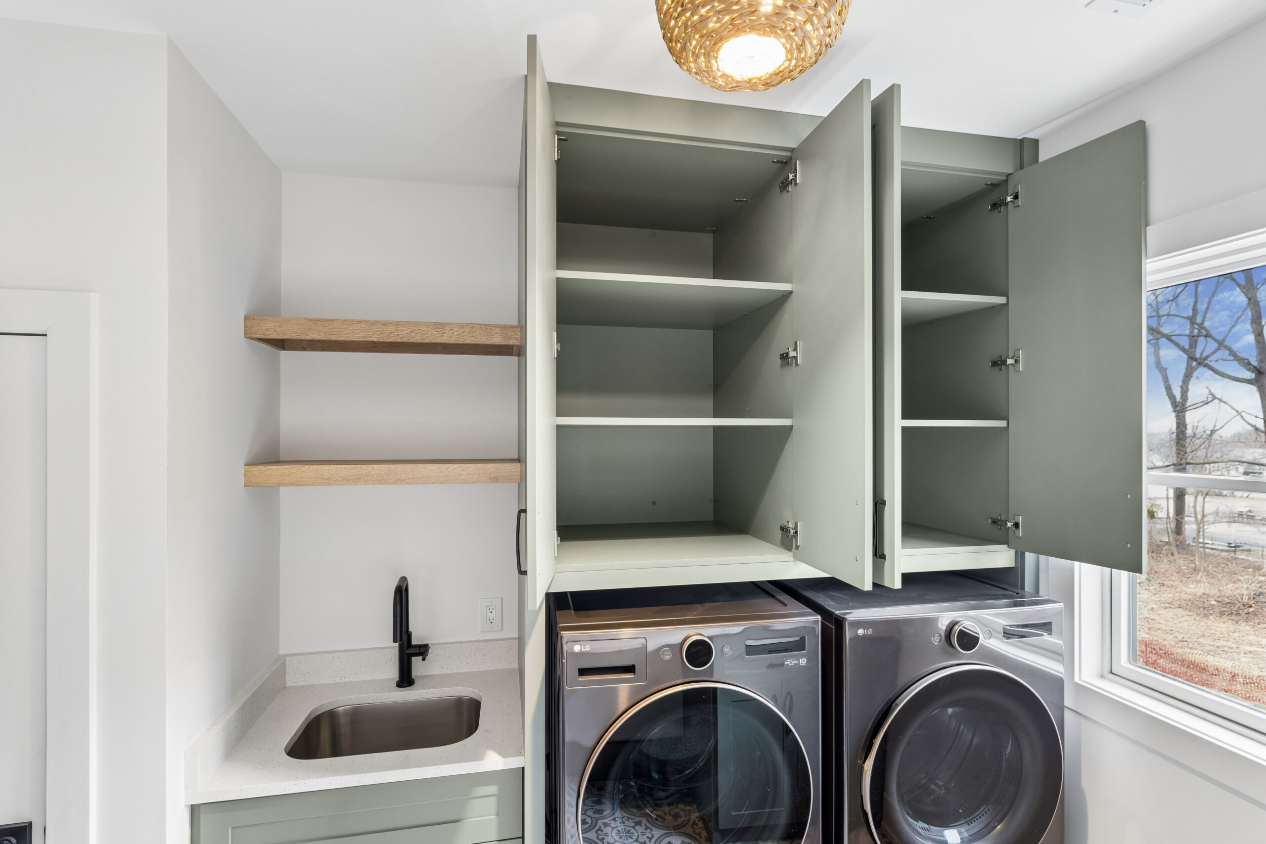 Modern laundry room with light green cabinets open to reveal empty shelves, a black sink, light wood floating shelves, and a front-loading washer and dryer beneath a window with a view outside.