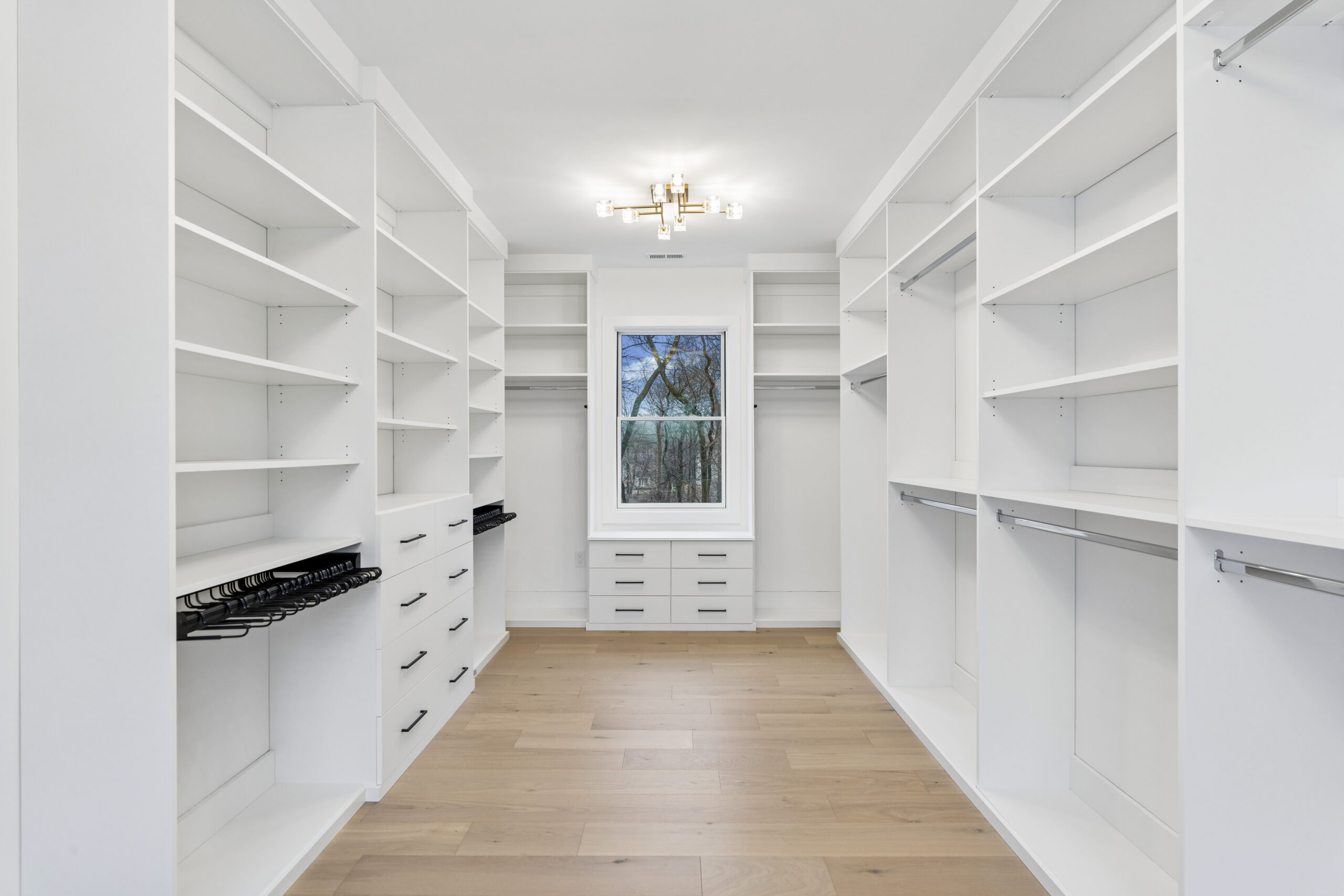 Spacious walk-in closet with white built-in shelves, drawers, and hanging rods on both sides, light wood flooring, a large window at the end, and a modern ceiling light fixture.