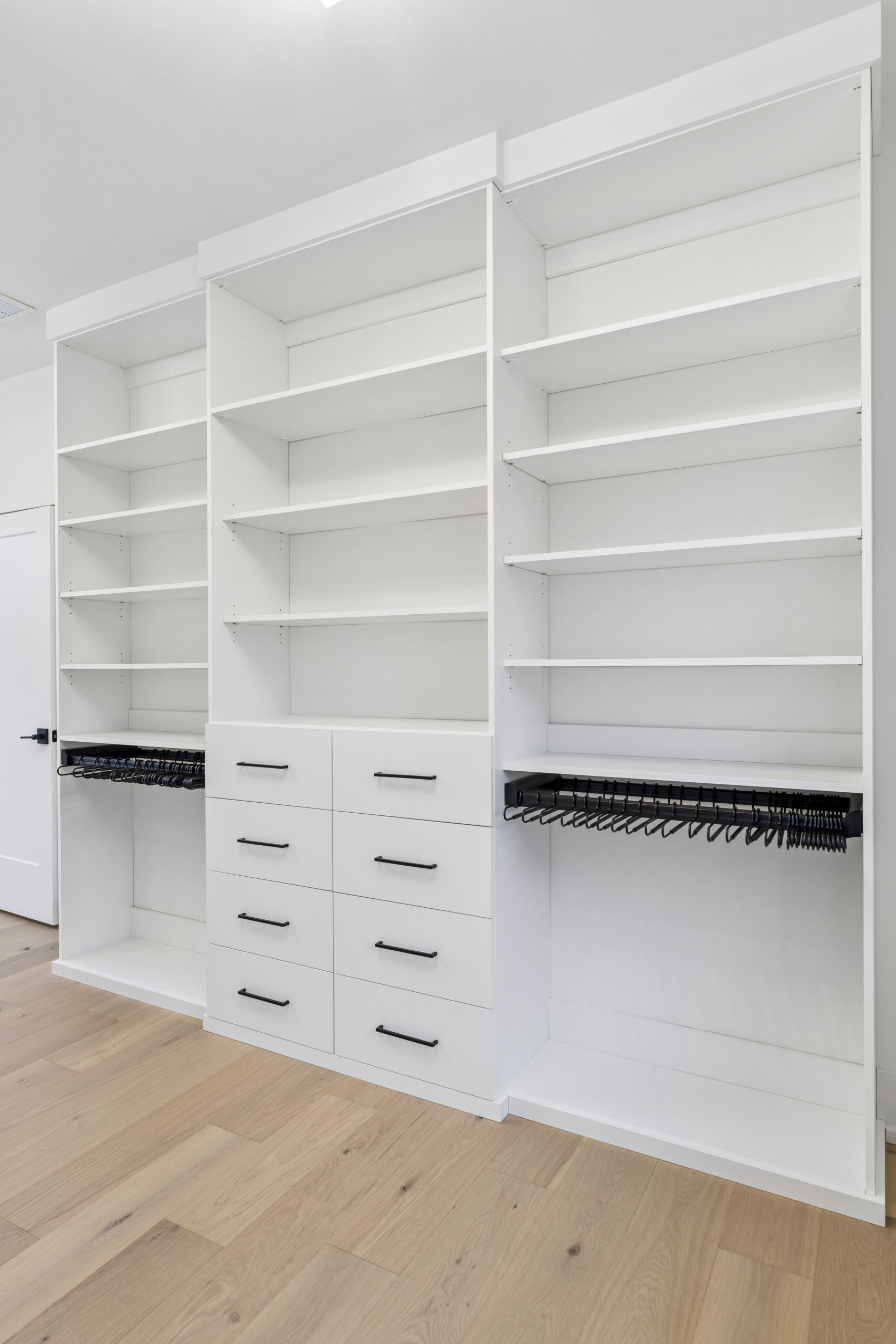 A modern closet with white shelves, multiple drawers with black handles, and black racks for hanging clothes, set against a light wood floor.
