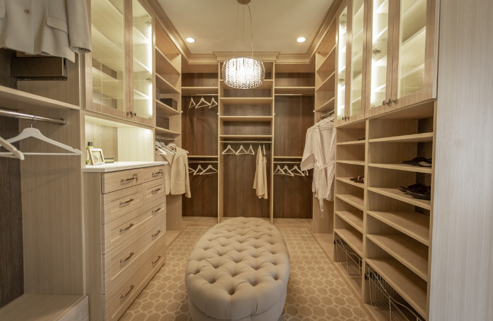 A large walk-in closet featuring light wood shelving, glass-front cabinets, hanging clothes, and a tufted fabric ottoman in the center.