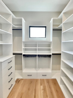 A walk-in closet with white shelving, clothing rods, and drawers, featuring a small rectangular window on the back wall.